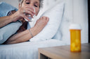 A middle aged woman laying in bed with arm resting on pillow holding a glass of water to her mouth. An orange pot of tablets sit on the bedside table