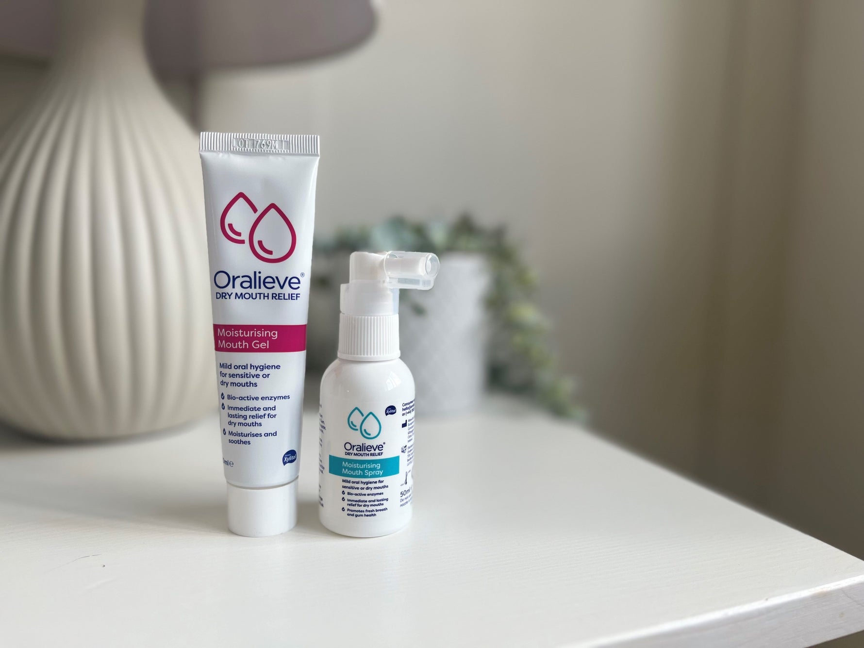 Oralieve spray and gel packs sitting on a white bedside table with a lamp and a trailing plant in pot in the background