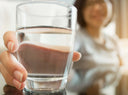 A large glass of water in focus being held by a women in the background which is blurred